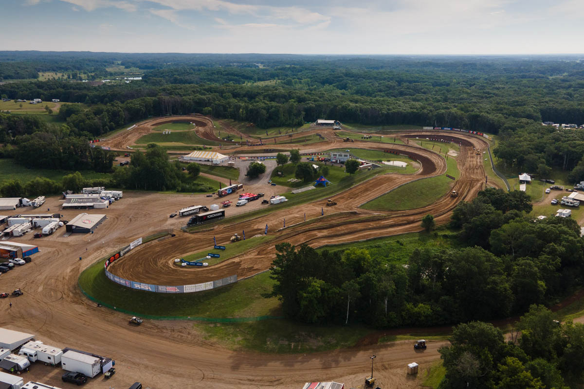 An aerial view shows an outdoor racing event grounds, featuring a large parking and pit area with numerous vehicles and trailers near a small pond.