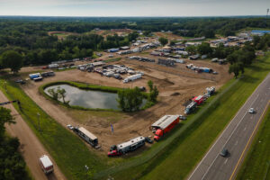 An aerial view shows an outdoor racing event grounds, featuring a large parking and pit area with numerous vehicles and trailers near a small pond.