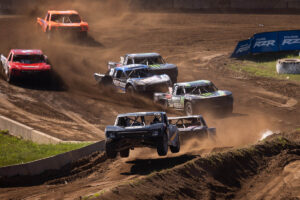 Multiple off-road racing trucks navigate a dirt track, with one truck airborne over a jump in the foreground.