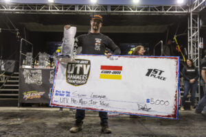 Scott LeSage on the KOH podium holding a giant fake check and a trophy while dawning a Maxxis podium hat.
