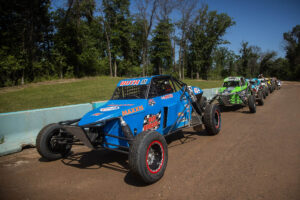 A line of off-road buggies, led by a prominent blue vehicle, is staged on a dirt track, likely preparing for or returning from a race.