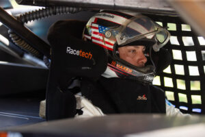 A race car driver wearing a helmet sits inside the cockpit of a vehicle.