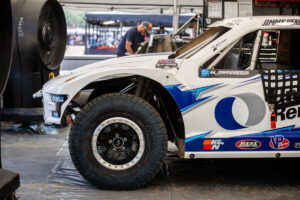 A white off-road racing truck sits in a garage area, with a mechanic working on a vehicle in the background.