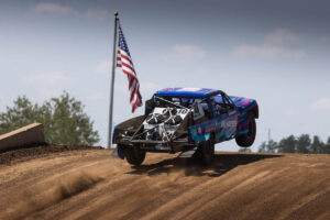 A blue and purple racing truck jumping over a dirt mound, with an American flag flying in the background.