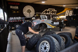 A mechanic works on a racing truck inside a garage filled with spare tires and team branding.