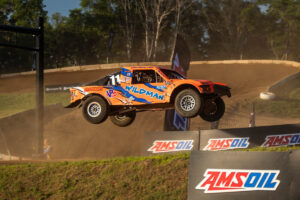 An orange off-road racing truck jumps through the air over an AMSOIL-branded dirt track.