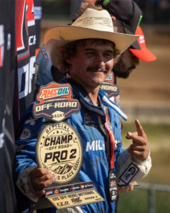 A racing driver wearing a cowboy hat holds a Pro 2 second-place trophy and makes a hand gesture.