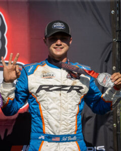 A racing driver wearing a white and blue suit holds up three fingers while wearing a race medal.