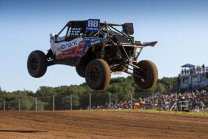 A racing truck leaps through the air over a dirt track with spectators in the background.