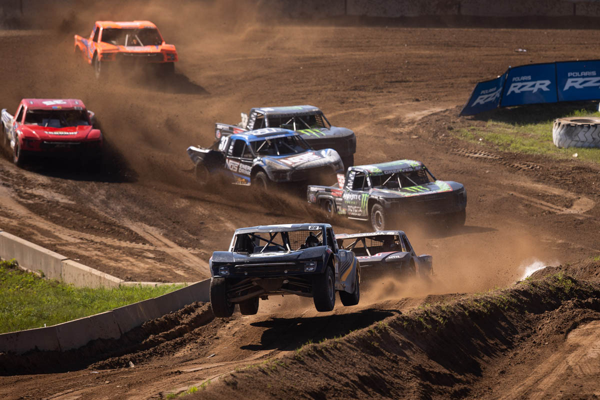 Multiple off-road racing trucks navigate a dirt track, with one truck airborne over a jump in the foreground.