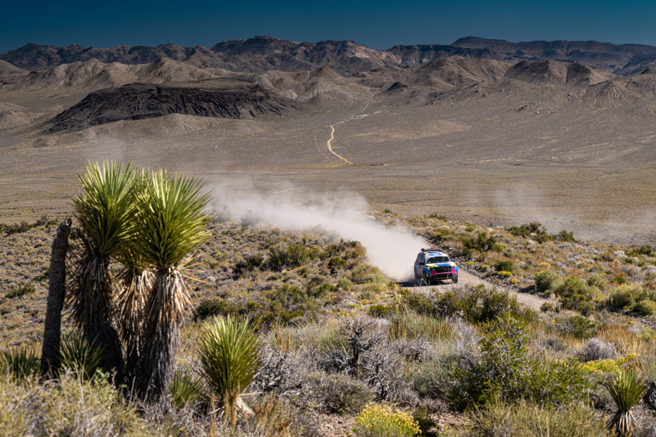 Scenic action image of Team #112 Honda Passport TrailSport, Rebelle Rally vehicle racing through dirt road in off-road terrain with mountains in the background.