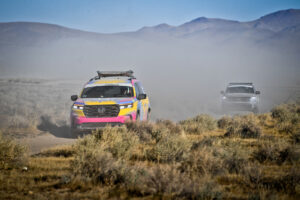 Team #211 Honda Pilot Trail Sport vehicle leading ahead of another other vehicle on dirt road with mountains in the background.