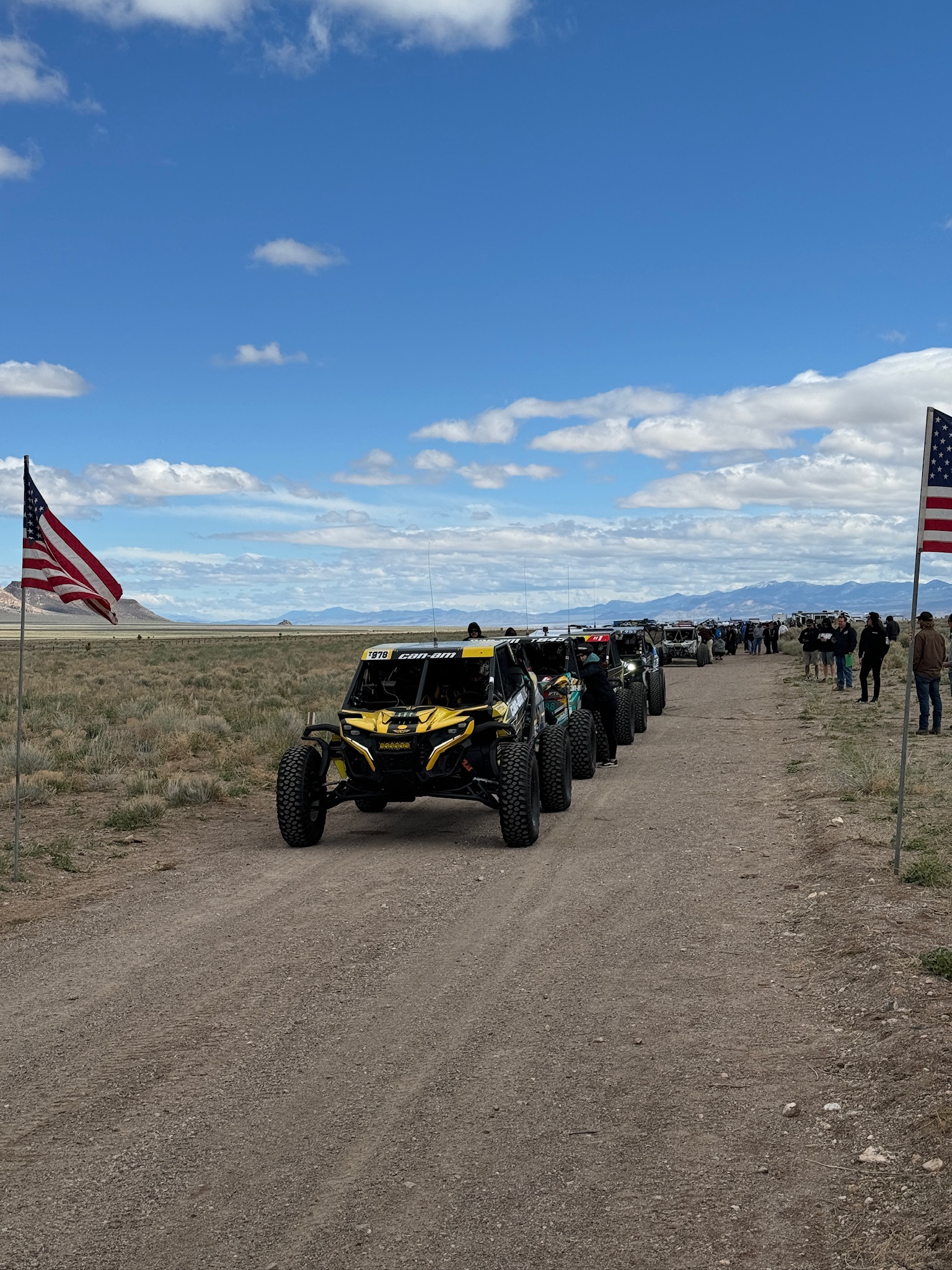 A line of SxS sitting on a dirt road staged for the start of the Silver State 300.