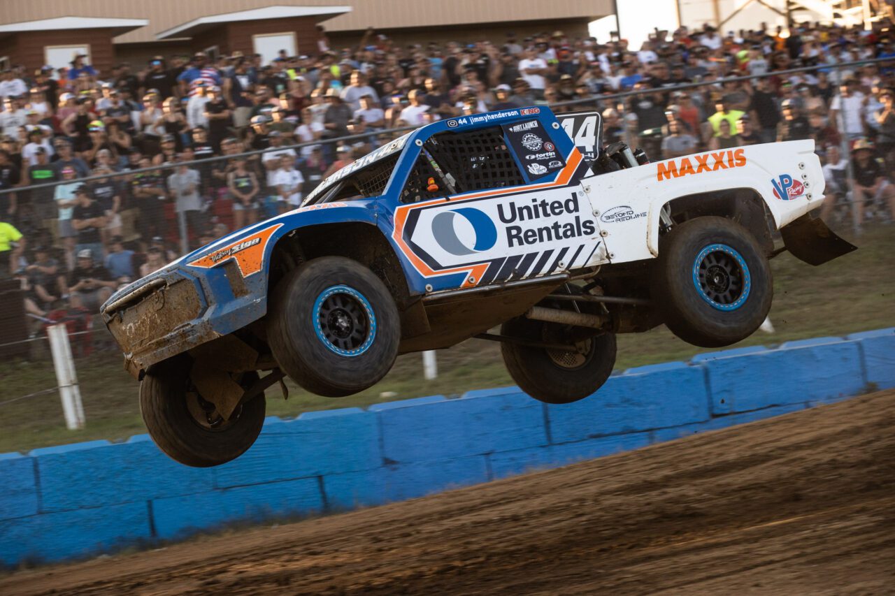 White and Blue truck airborn in front of fans standing on a hill in the background.