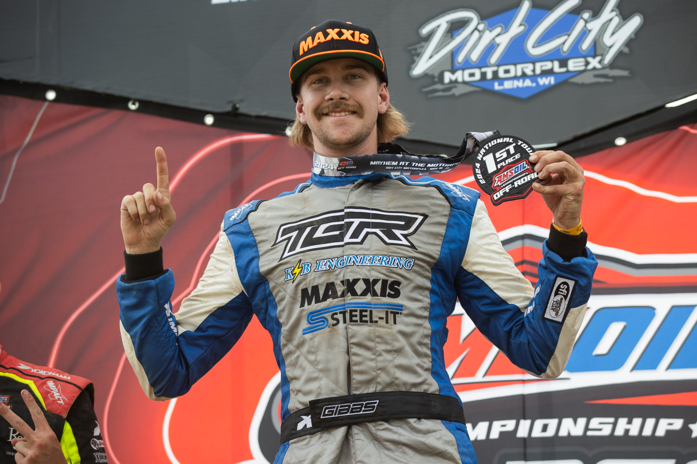 Trey Gibbs celebrating on the podium ar Round 2. Holding up a number one finger with one hand and his 1st place medal with the other hand.