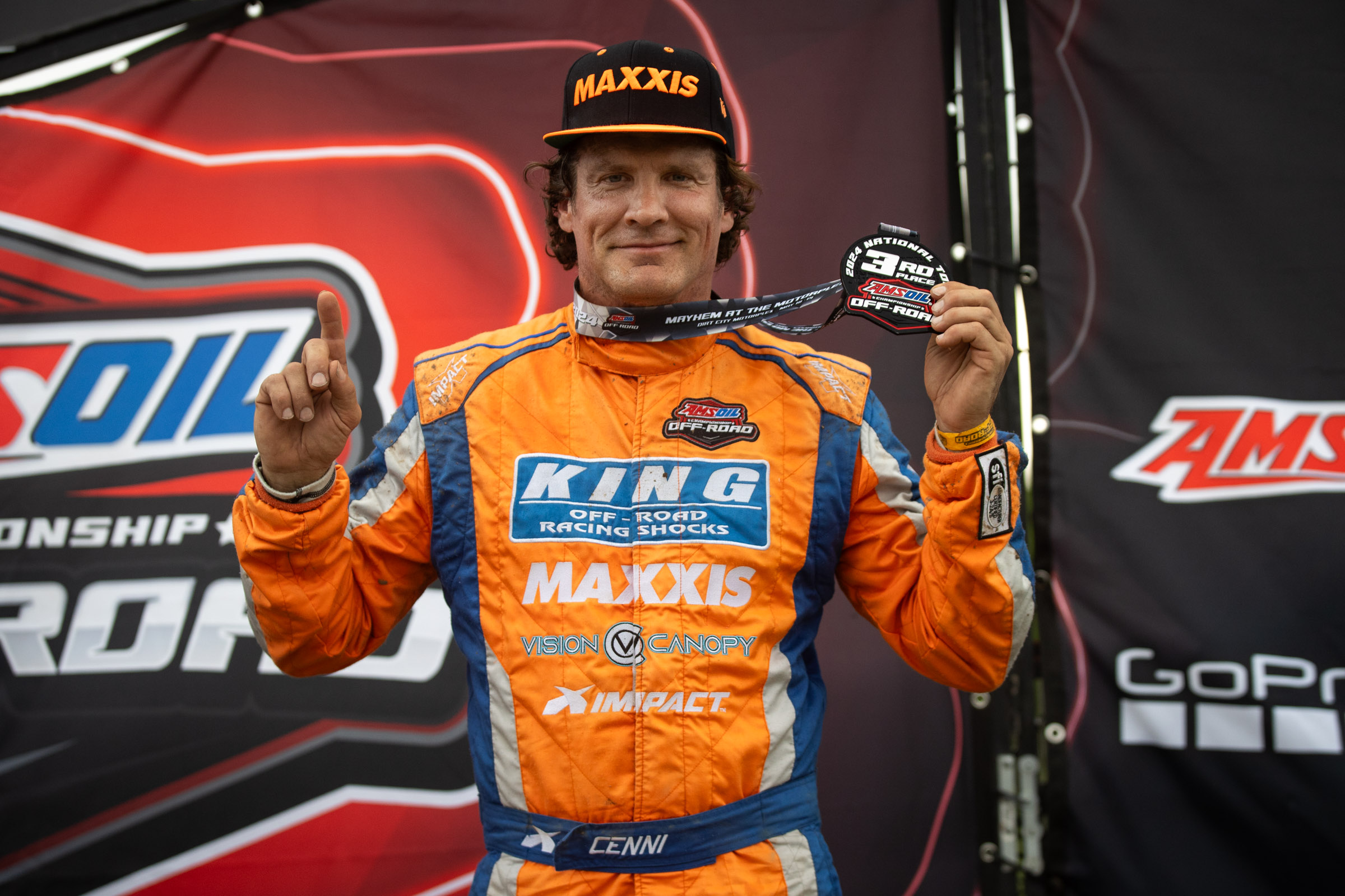 Cenni standing on the podium holding a first place medal with his number one finger held up and wearing a maxxis hat.