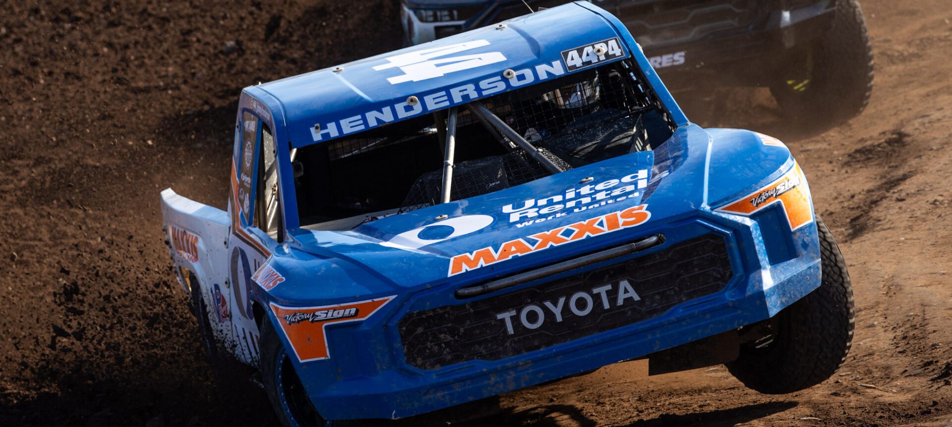 Blue and orange race truck leading the race out of a a dirt bowl turn.