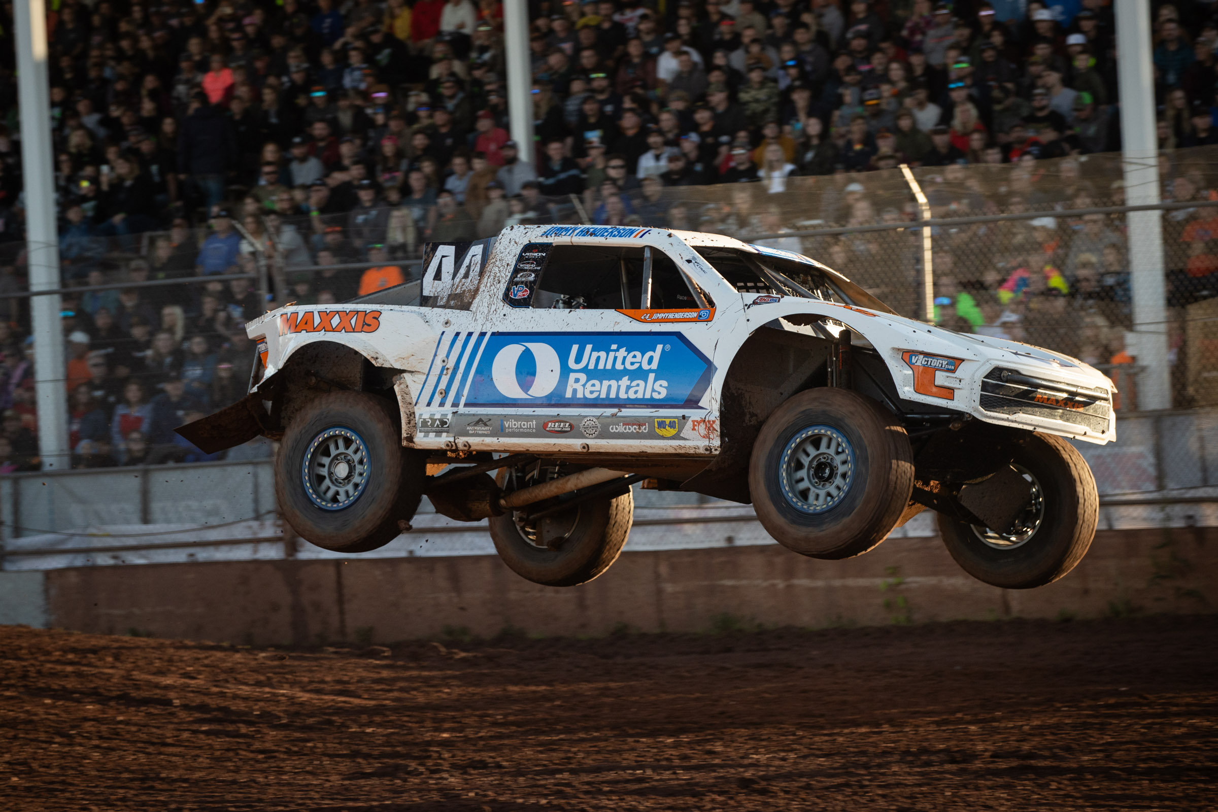 Jimmy Henderson in his white/blue/orange Pro4 race truck in mid flight and about to touch down. The grand stands in the background are full of race fans.