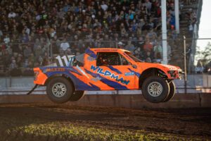 Cenni in his fluorescent orange and blue race truck in mid flight. The grand stands in the background are full of race fans.