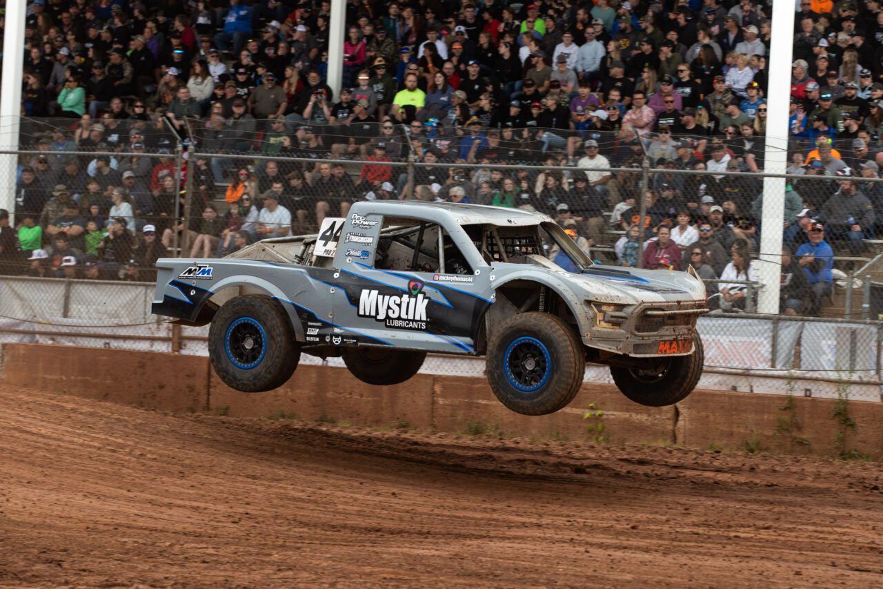 Thomas in his Mystick grey/blue/black Pro2 race truck in mid flight. The grand stands in the background are full of race fans.