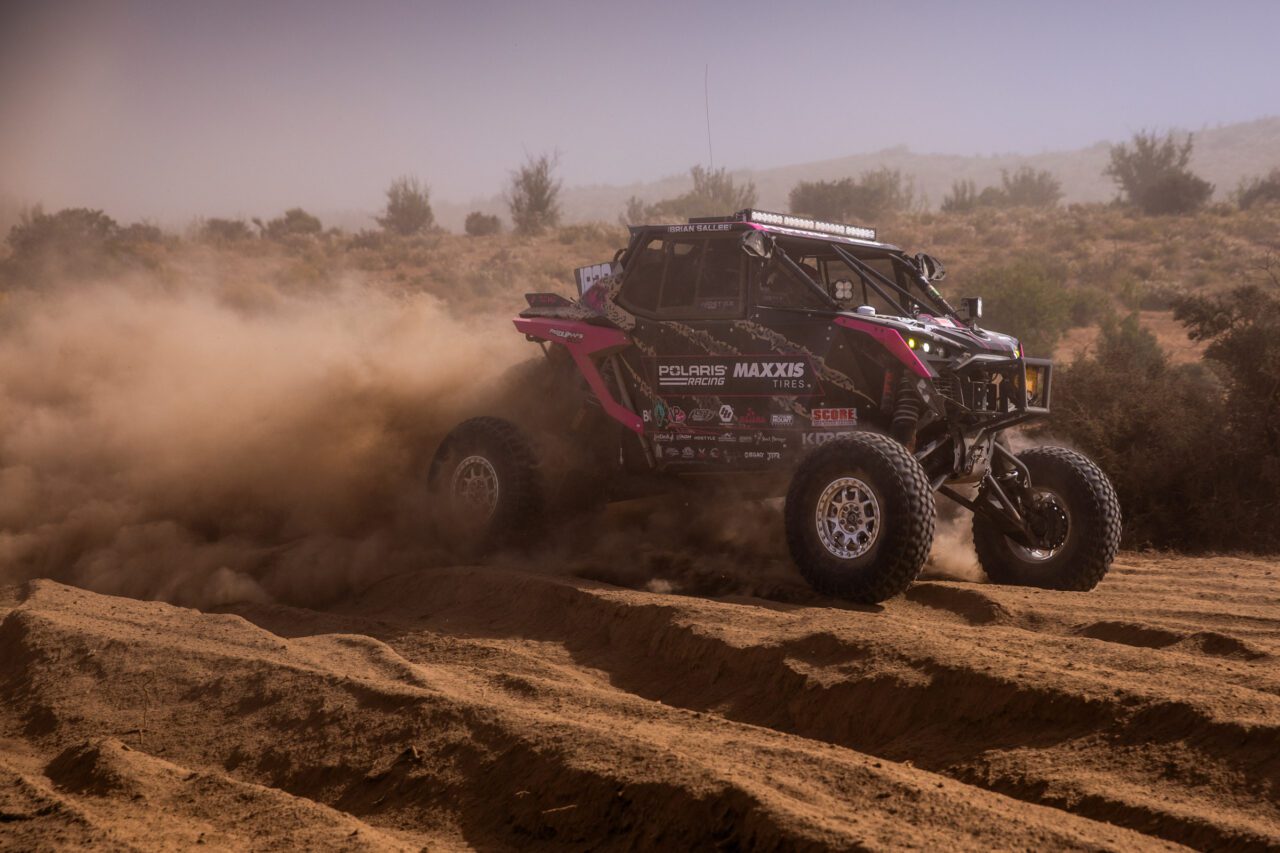 Pink and black SxS at full speed in the desert with large plume of dust trailing behind it.