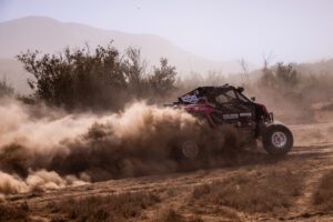Pink and black SxS at full speed in the desert with large plume of dust trailing behind it.