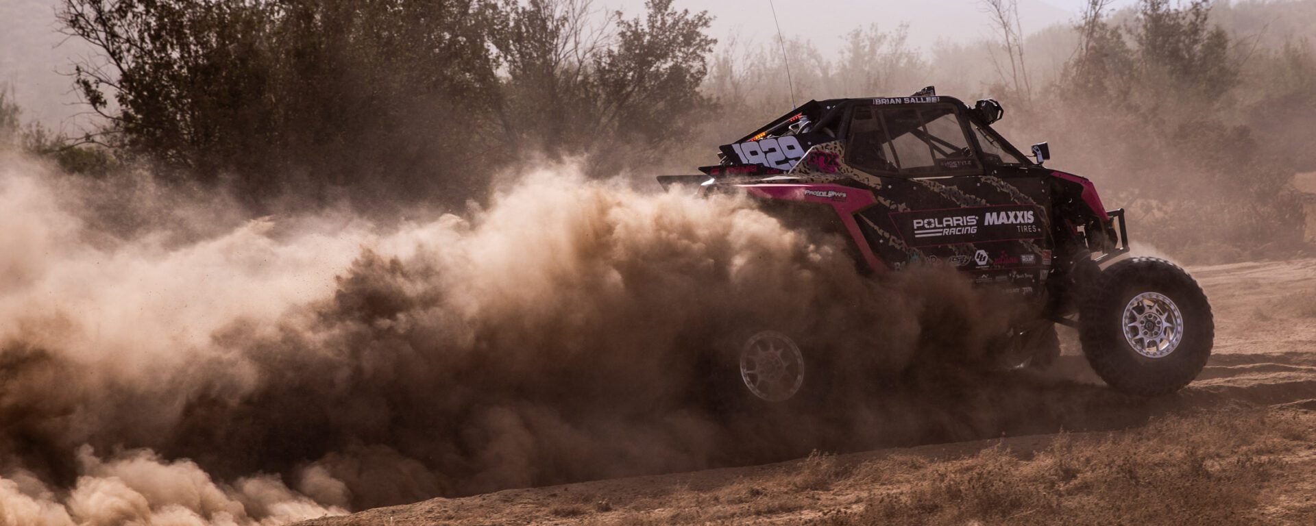 Pink and black SxS at full speed in the desert with large plume of dust trailing behind it.