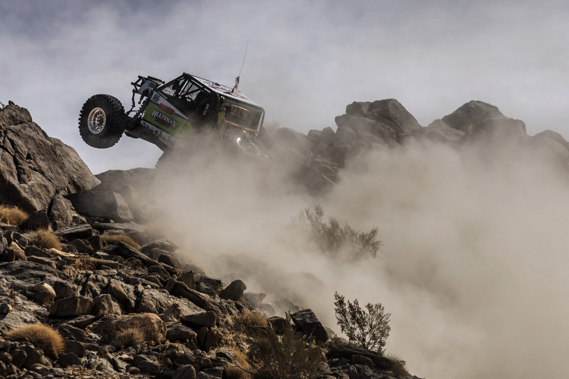Offroad Truck, going up a hill. One tire lifted in the air with a large dust cloud behind.