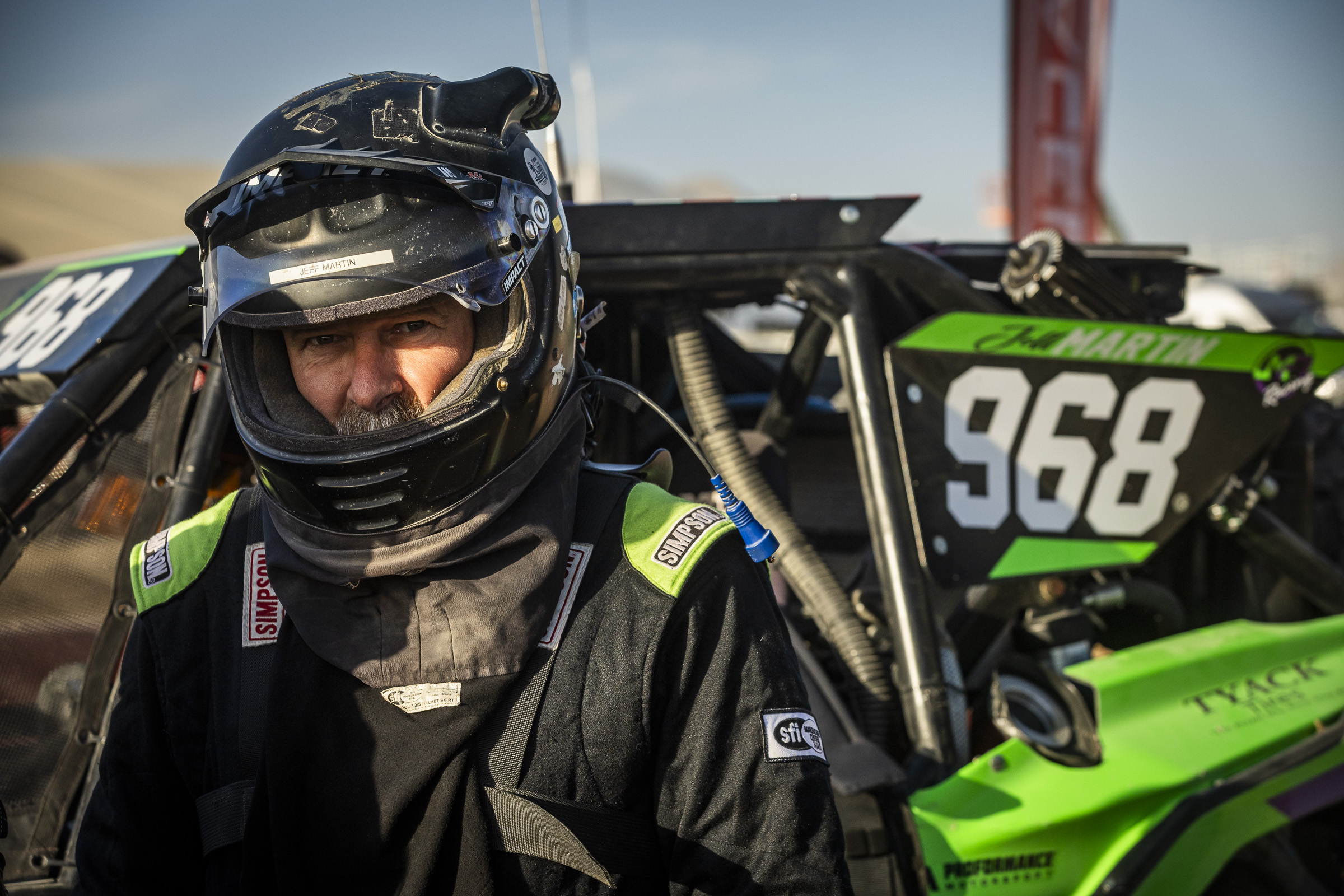 Un pilote casqué se tenant devant sa voiture de course. Le numéro de course 964 est bien visible sur le côté de la voiture en arrière-plan.