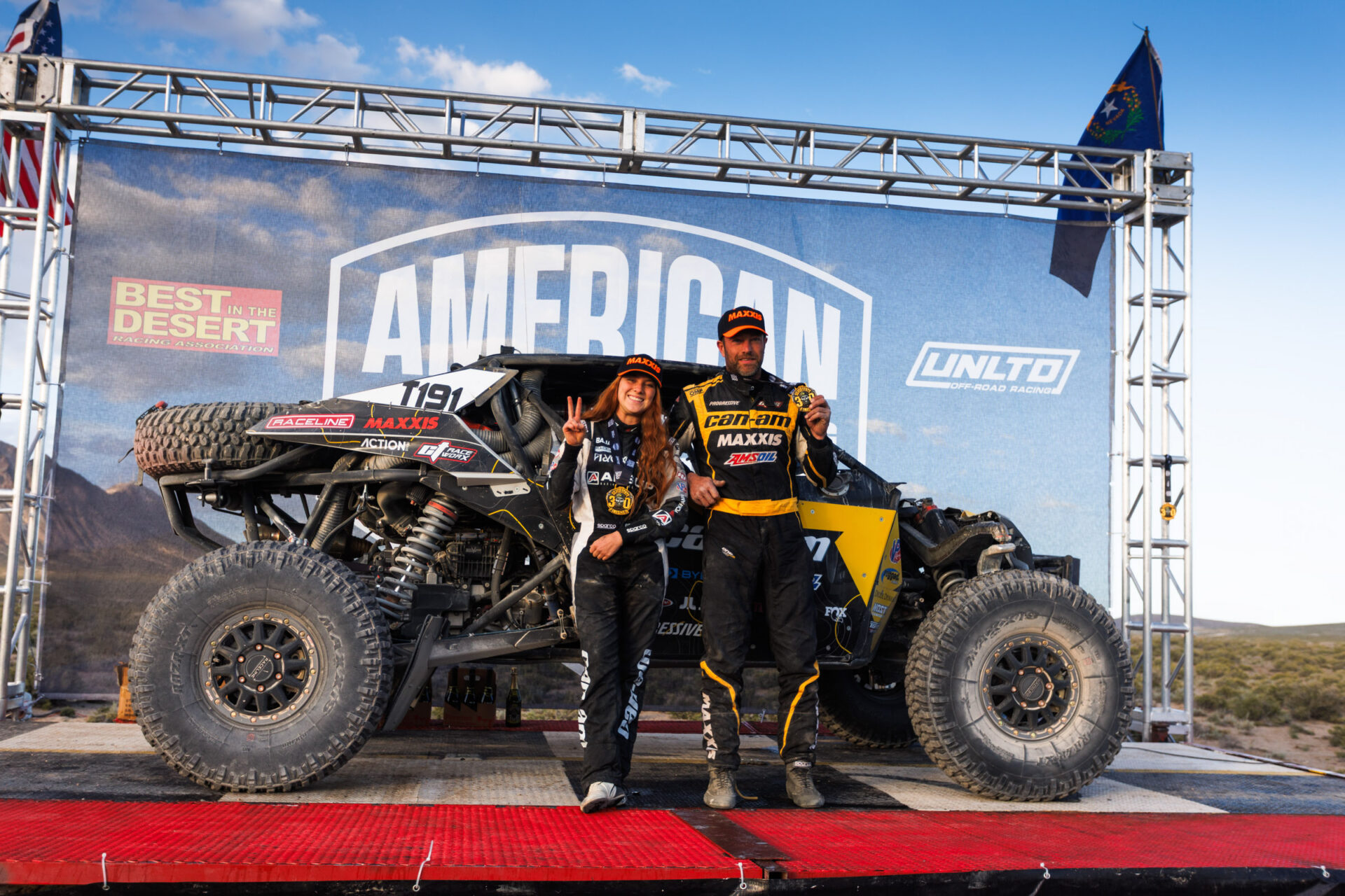 Kyle Chaney and his co-driver in black and yellow driving suits standing on the BITD poduim in front of their race car holding up number one for first place.