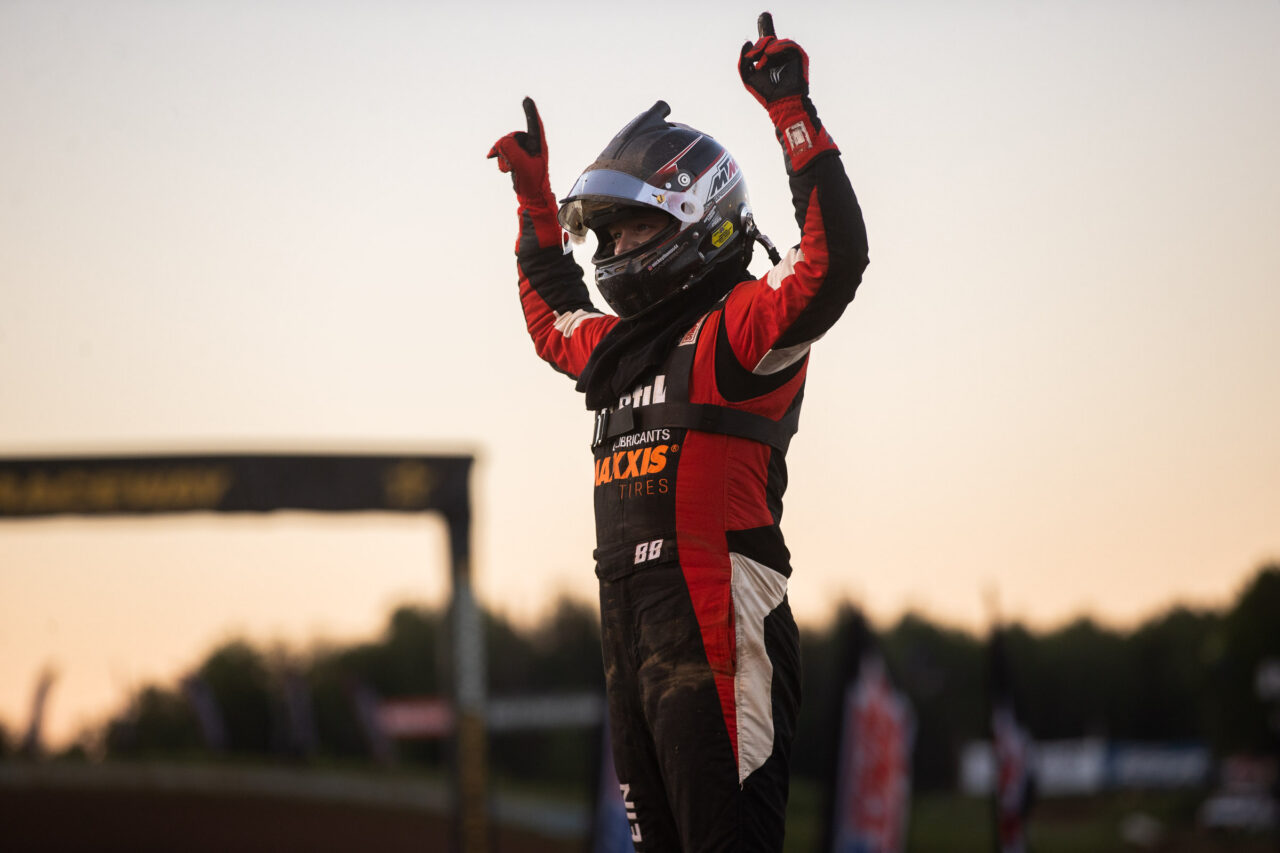 a driver in full fire suit and helmet with both hands raised holding up number one after a hard earned win on Maxxis tires.