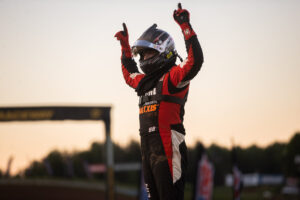 a driver in full fire suit and helmet with both hands raised holding up number one after a hard earned win on Maxxis tires.