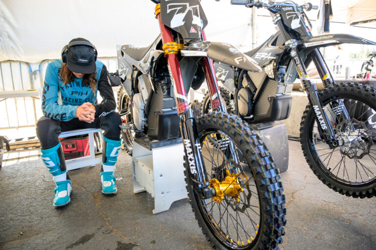 Pat Evans sitting looking downward. Headphones on and sitting next to his dirt bike. The image looks like he is mentally preparing for his ride.