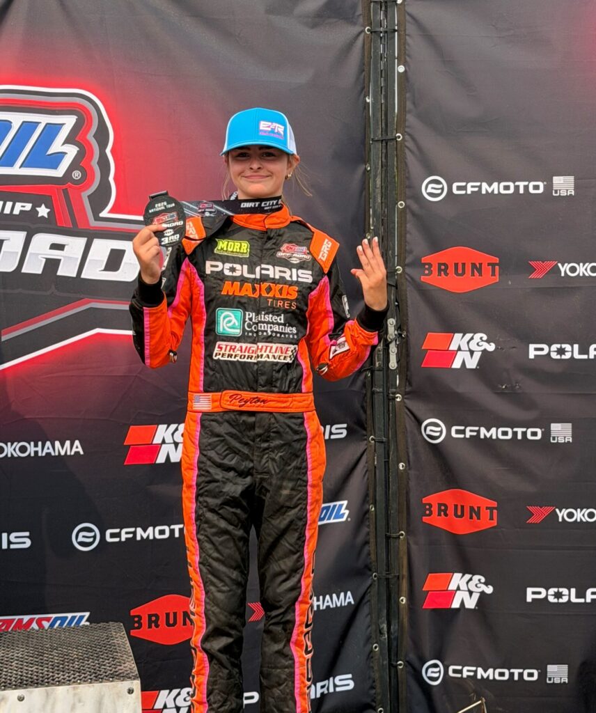A driver wearing a blue hat and an orange, black, and pink racing suit stands proudly on the podium, holding a 3rd-place medal in one hand while making a three-finger gesture with the other.