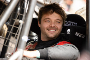 Smiling driver sitting inside the safety netting and bucket seat of a race car.