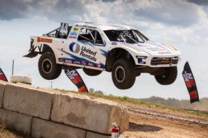 White United Rentals pro-4 off-road truck airborne as it leaps over a dirt ramp.
