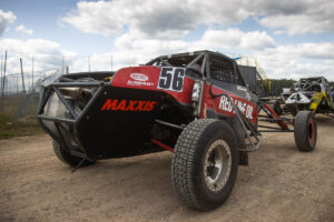 Red number 56 off-road racing buggy parked outdoors on a dirt track under a partly cloudy sky.