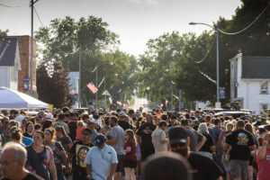 Large crowd of people walking down a tree-lined main street during an outdoor community festival.