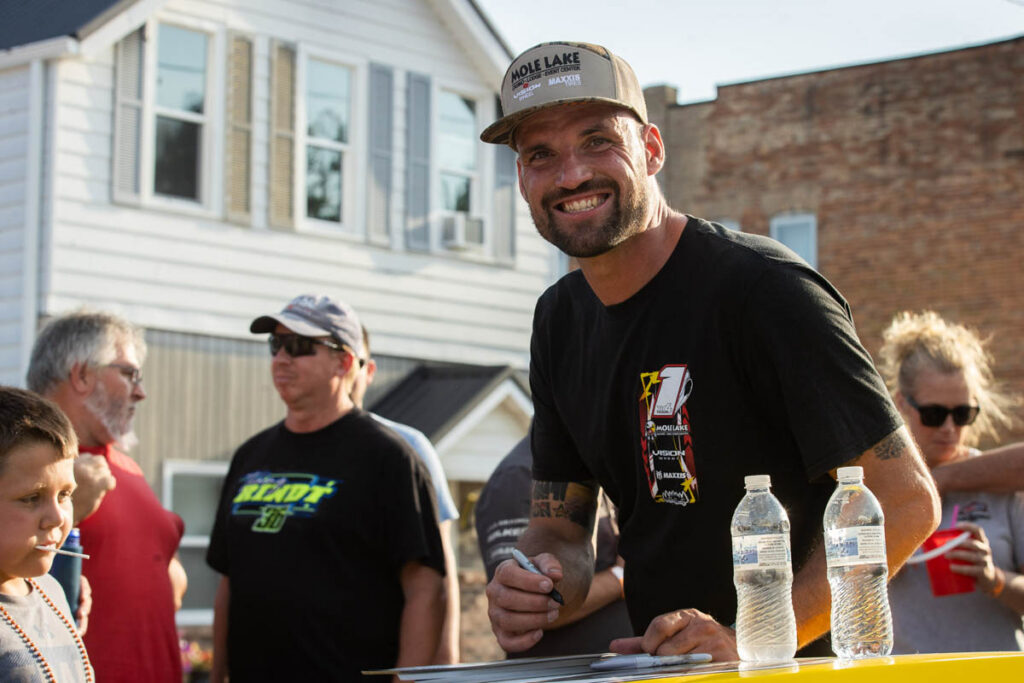 Smiling driver wearing a cap signing autographs for fans at an outdoor event.