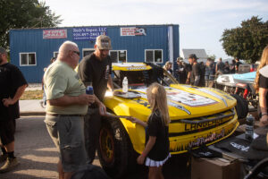 Driver in a hat interacting and handing items to fans next to a yellow off-road race truck.