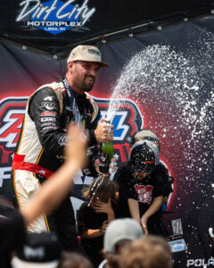 A driver celebrates on the podium, spraying champagne over fans and children nearby.