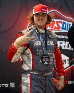 Zach Drapkin smiles on the podium flashing a peace sign with a 2nd-place medal.