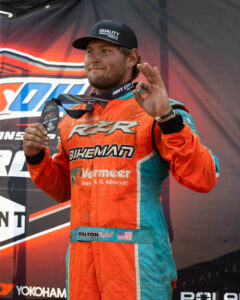 Kolton Krajicek shows an OK sign while holding a 3rd-place medal in his racing suit.