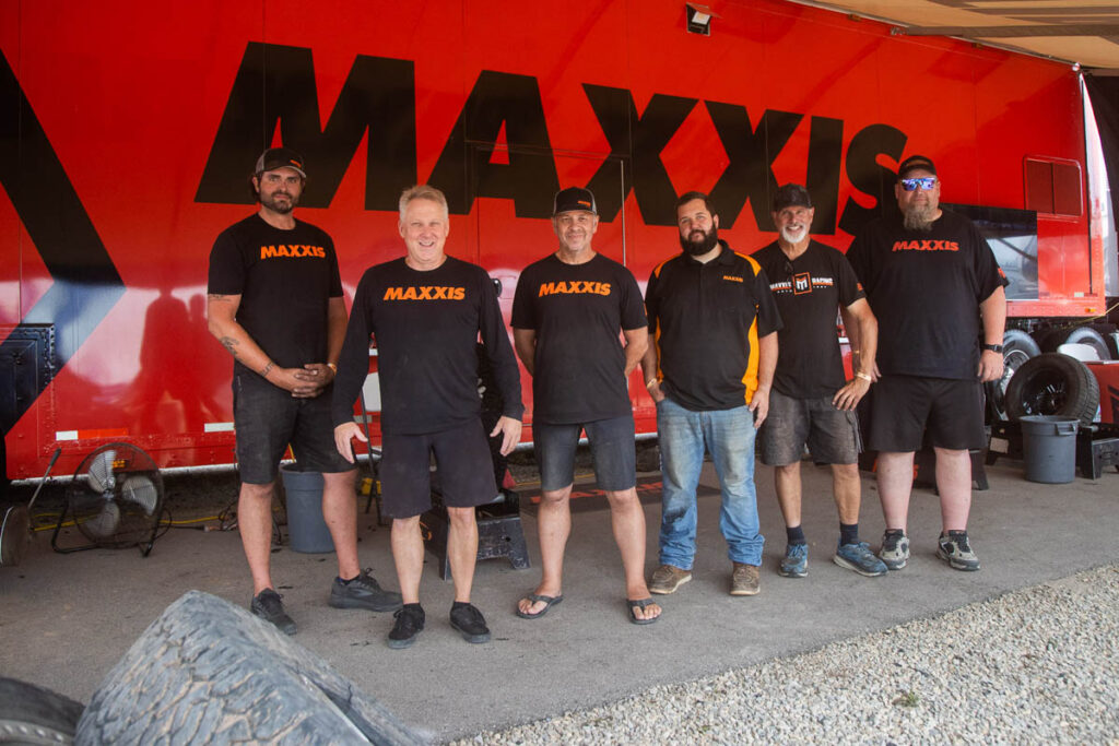 A group of six team members wearing Maxxis apparel pose together in front of a red Maxxis team transport truck.