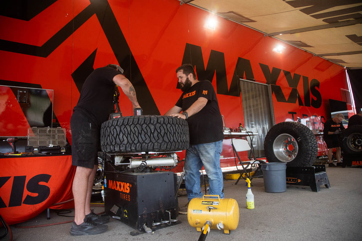 Two mechanics working on a heavy-duty off-road tire inside a garage or pit tent with Maxxis branding.