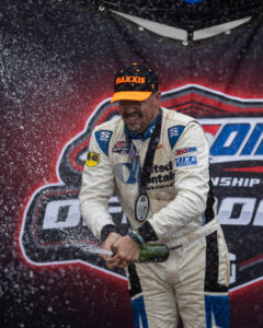 Racing driver in a white and blue suit spraying champagne on the podium amidst a shower of spray.