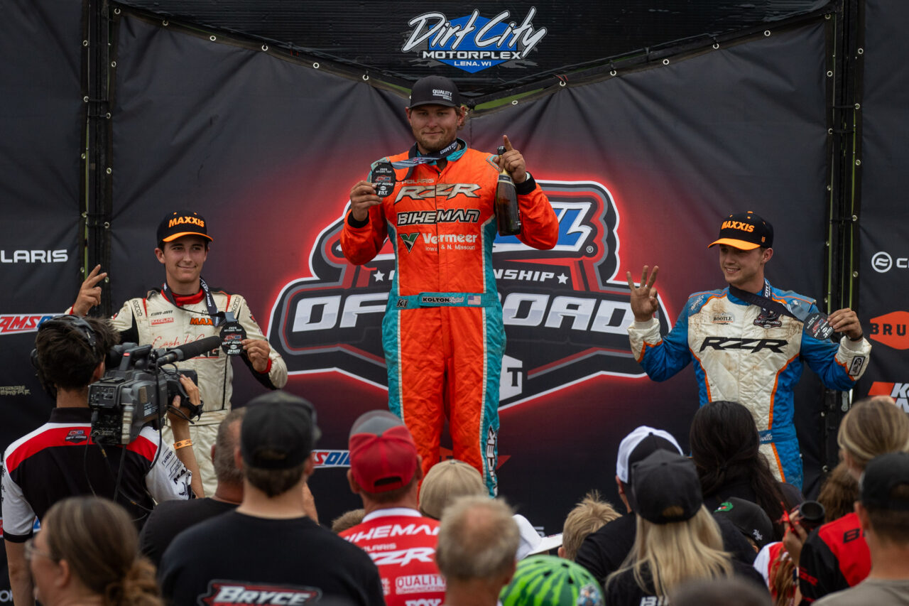 Three off-road racing drivers on a podium celebrating after a race, holding medals and a champagne bottle, with a crowd and media crew watching from below in front of a Dirt City Motorplex backdrop.
