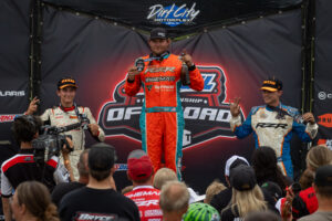 Three off-road racing drivers on a podium celebrating after a race, holding medals and a champagne bottle, with a crowd and media crew watching from below in front of a Dirt City Motorplex backdrop.