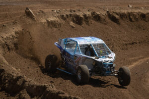 A white and blue off-road vehicle driving around a dirt corner, kicking up dust and dirt from the track.