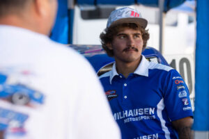 A driver wearing a Milhausen Motorsports shirt and a baseball cap talks to someone in a pit area.A driver wearing a Milhausen Motorsports shirt and a baseball cap talks to someone in a pit area.